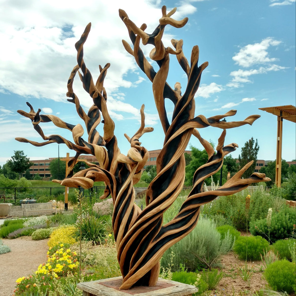 A large twisted juniper wooden sculpture resembling a stylized tree or natural form, mounted on a square pedestal in an outdoor garden setting