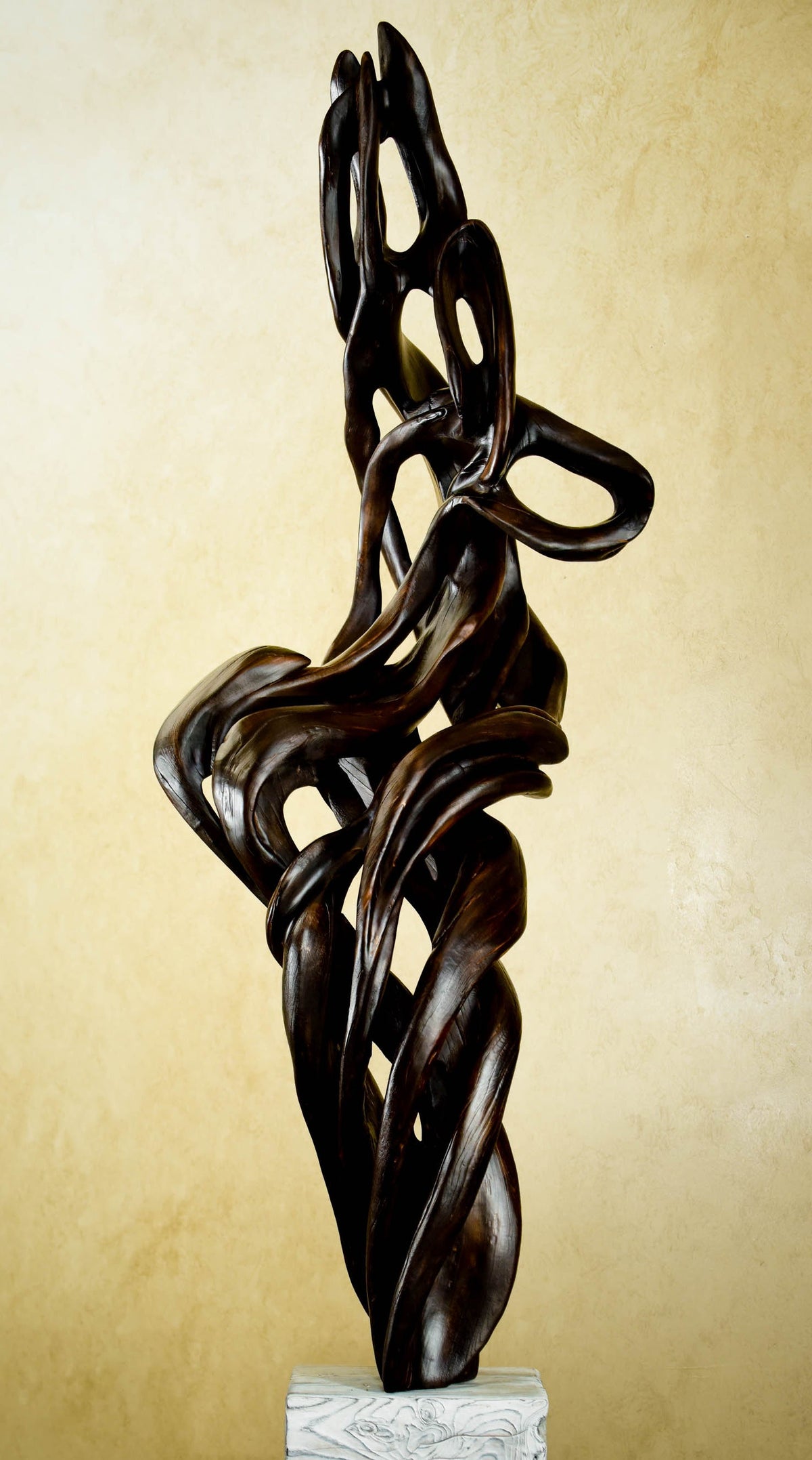 An abstract dark ebony colored  juniper wood sculpture with flowing, intertwined organic curves, displayed on a white block base against a textured beige background.