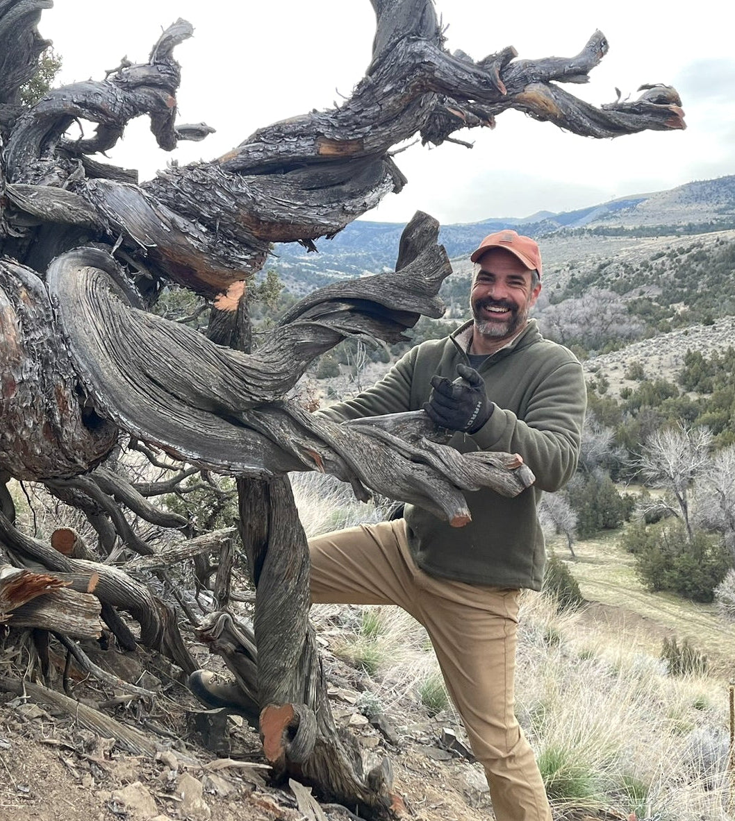 Tom Benedict stands next to a large, gnarled ancient tree in a rugged, rocky outdoor landscape with hills in the background.