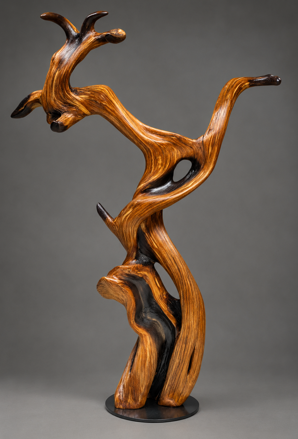 A sculptural wooden tree sculpture with dramatic curved branches and natural grain, displayed on a round steel base against a neutral gray studio background. torched wood ends.
